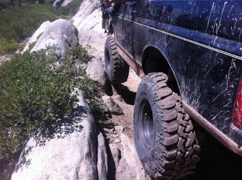 1990 Ford Bronco - A dark-colored off-road vehicle is shown from the side, driving over a rocky path with vegetation.