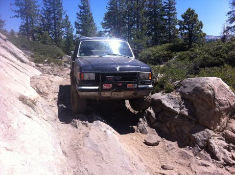 1990 Ford Bronco - A black Ford Bronco tackles a challenging off-road path with large rocks and trees.