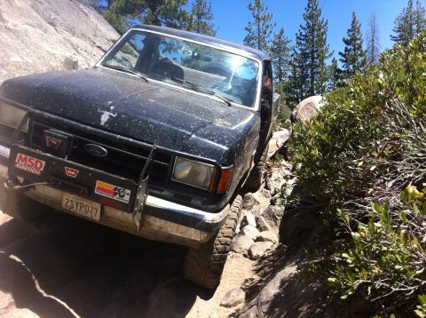 1990 Ford Bronco - A black Ford pickup truck navigates a challenging rocky trail with dense foliage on the side.