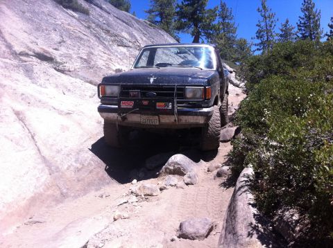 1990 Ford Bronco - A black Ford Bronco navigates a challenging off-road trail with rocky terrain and dense vegetation.