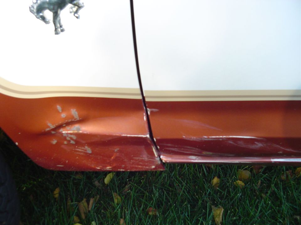 Dented, scraped copper lower body panel below white paint with a chrome bucking-horse fender emblem