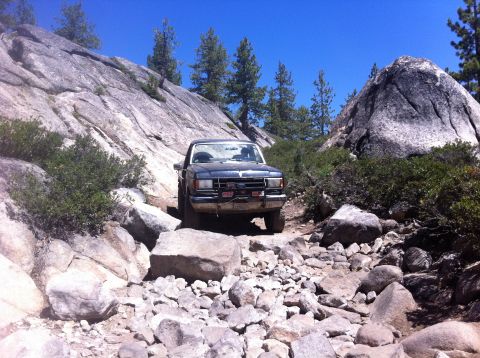 1990 Ford Bronco - A Ford Bronco tackles a challenging rocky terrain on a sunny day, surrounded by boulders and sparse vegetation.
