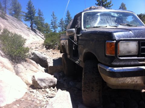1990 Ford Bronco - A black Ford Bronco navigates a challenging rocky trail under a clear blue sky.