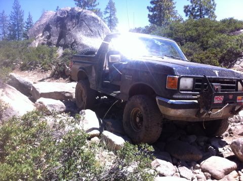 1990 Ford Bronco - This 1990 Ford Bronco is shown tackling a challenging off-road course with large rocks and dense vegetation.