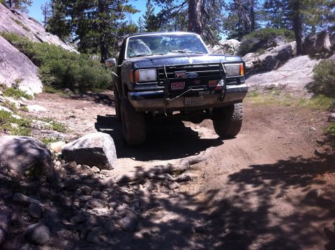 1990 Ford Bronco - 1990 Ford Bronco navigating a rough, rocky trail in a wooded, mountainous area.