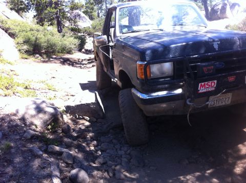 1990 Ford Bronco - This truck is navigating a rough, rocky off-road path with trees and boulders visible.