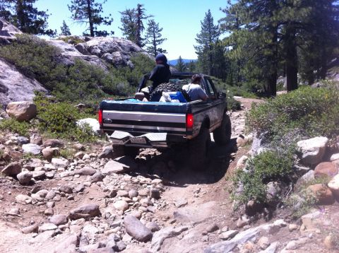 1990 Ford Bronco - 1990 Ford Bronco navigates a challenging off-road path in a wooded, rocky area.