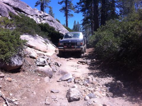 1990 Ford Bronco - A dark-colored pickup truck navigates a challenging, boulder-strewn path through a wooded, mountainous area.