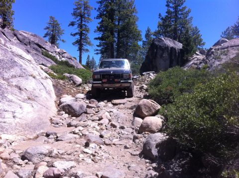 1990 Ford Bronco - This SUV navigates a challenging, rocky path through a wooded, mountainous area under a clear blue sky.
