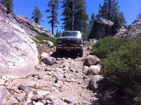 1990 Ford Bronco - A black Ford Bronco navigates a challenging rocky path in a mountainous, wooded area under a clear blue sky.