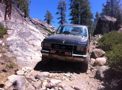 1990 Ford Bronco - A black Ford Bronco navigates a challenging, rocky trail under a clear blue sky.