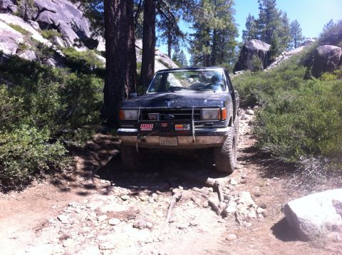 1990 Ford Bronco - A dark-colored Ford Bronco navigates a rough, off-road path surrounded by trees and large rocks.