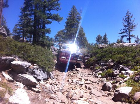 1990 Ford Bronco - A Ford Bronco drives through a challenging, rocky terrain surrounded by trees and boulders.