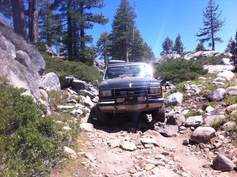 1990 Ford Bronco - A black Ford Bronco navigates a challenging, rocky off-road trail under a clear blue sky.