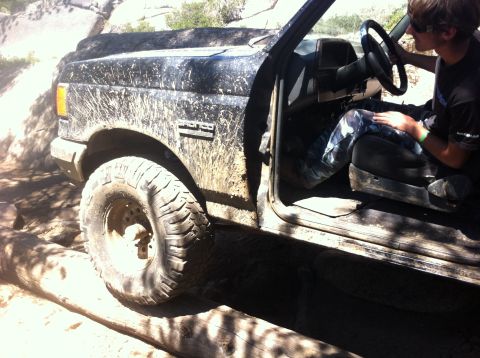 1990 Ford Bronco - A muddy 1990 Ford Bronco navigates a rocky trail with logs in the foreground.