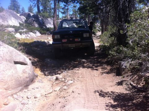 1990 Ford Bronco - A dark SUV navigates a challenging off-road trail with large rocks and trees.