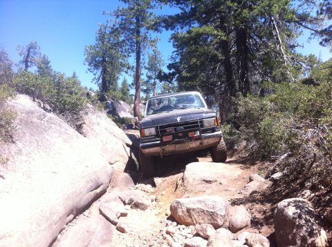 1990 Ford Bronco - 1990 Ford Bronco tackling a challenging off-road path in a wooded, rocky area.
