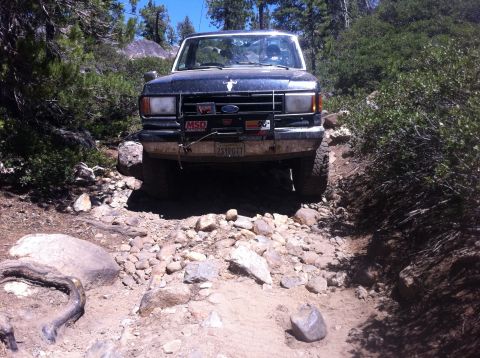 1990 Ford Bronco - A Ford Bronco navigates a challenging, rocky off-road path surrounded by dense vegetation.