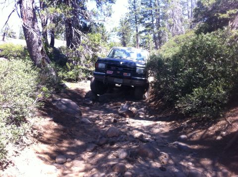 1990 Ford Bronco - A black SUV drives through a challenging, rocky off-road path surrounded by trees.