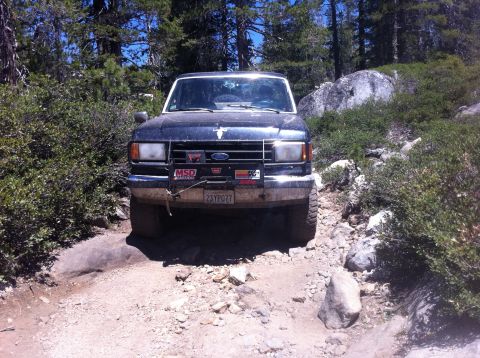 1990 Ford Bronco - A dark Ford Bronco navigates a rocky, off-road trail surrounded by trees and brush.