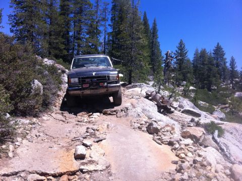 1990 Ford Bronco - A dark blue Ford Bronco navigates a challenging rocky trail in a wooded area.