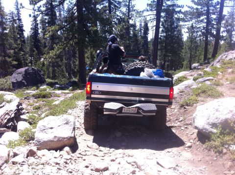 1990 Ford Bronco - A Ford pickup truck navigates a rocky, wooded trail with a spare tire loaded in the bed.