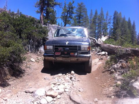 1990 Ford Bronco - A dark-colored Ford Bronco navigates a challenging, rocky path through a wooded, mountainous area.