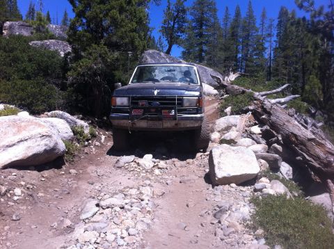 1990 Ford Bronco - A dark blue 1990 Ford Bronco navigates a challenging rocky trail surrounded by trees and boulders.