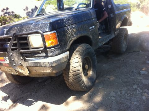 1990 Ford Bronco - A muddy black Ford Bronco navigates rocky terrain on an off-road trail.