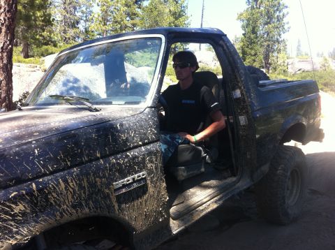1990 Ford Bronco - Mud-splattered 1990 Ford Bronco with driver visible through the open door.