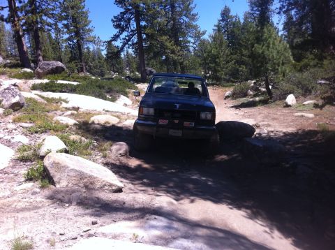 1990 Ford Bronco - A dark SUV navigates a rough, off-road trail surrounded by trees and boulders.