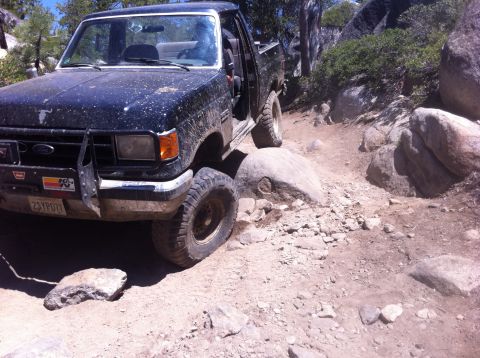 1990 Ford Bronco - A black Ford pickup truck navigates a rocky off-road trail, with one tire perched on a large boulder.