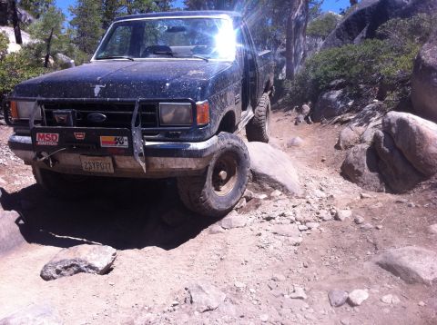 1990 Ford Bronco - A dark pickup truck maneuvers over large rocks on a dirt path.