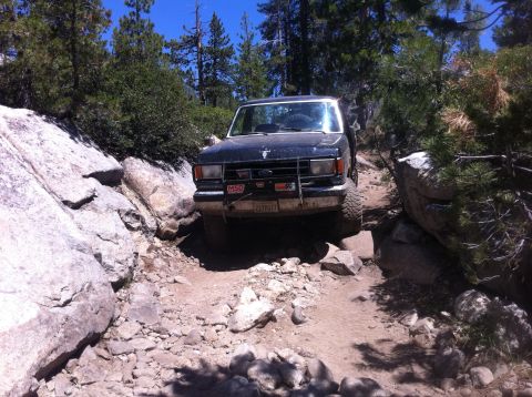 1990 Ford Bronco - This Ford Bronco II is tackling a challenging off-road path surrounded by rocks and pine trees.