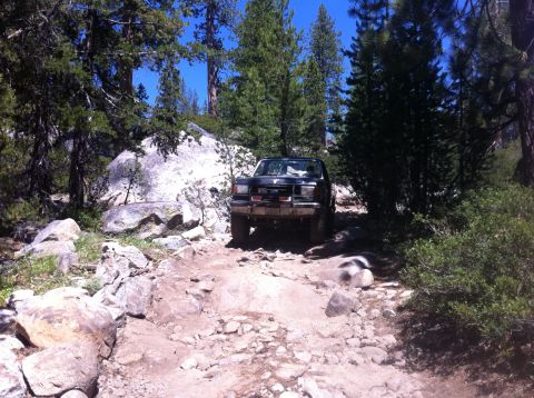 1990 Ford Bronco - A dark-colored truck is shown on a challenging, boulder-strewn path surrounded by trees and rocks.