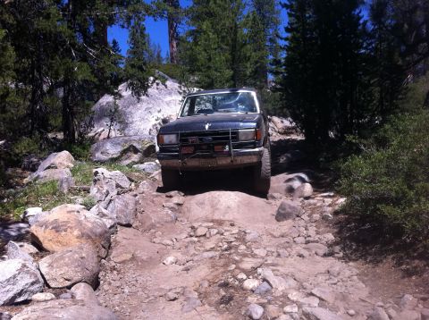 1990 Ford Bronco - A dark blue Ford Bronco navigates a challenging rocky terrain on a sunny day.