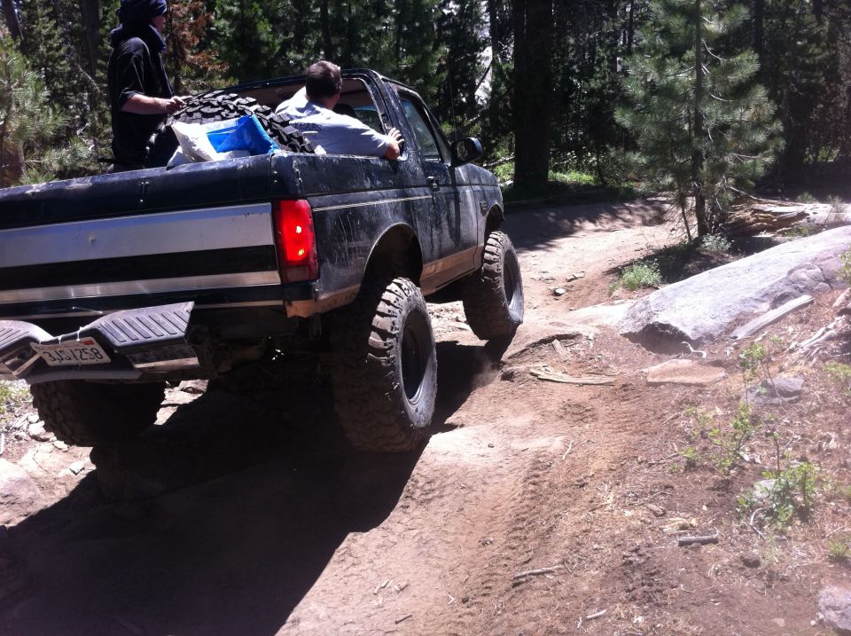 1990 Ford Bronco driving over rocks on a dirt trail, rear view. License plate reads JGS1258