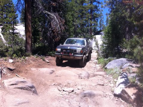1990 Ford Bronco - A black Ford Bronco navigates a challenging, rocky off-road trail surrounded by trees.