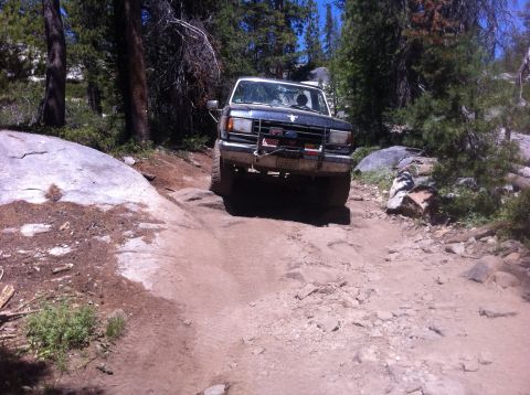 1990 Ford Bronco - A Ford Bronco navigates a rough, off-road path surrounded by trees and large rocks.