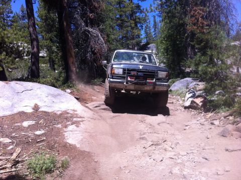 1990 Ford Bronco - A Ford Bronco navigates a challenging off-road path through a wooded, rocky area.