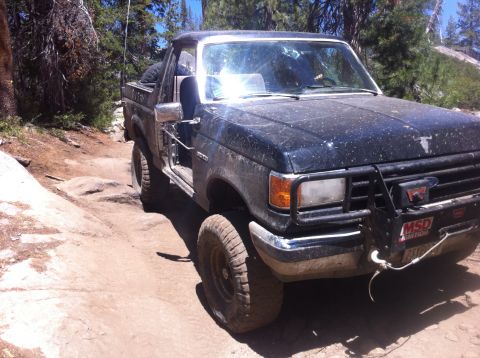 1990 Ford Bronco - This truck is navigating a rough, off-road path with trees and rocks visible.