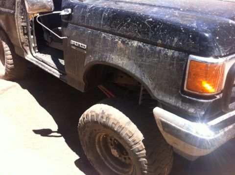 1990 Ford Bronco - Mud-covered Ford Bronco with the driver's door ajar, showing its front fender and tire.