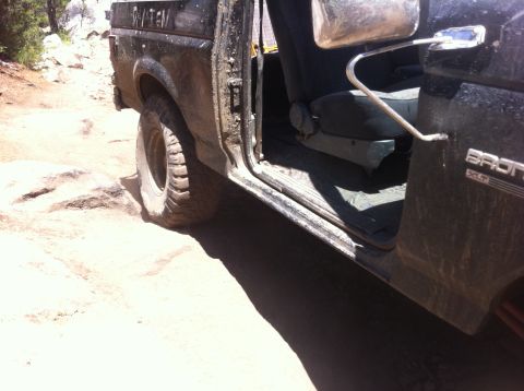 1990 Ford Bronco - This 1990 Ford Bronco is shown off-road, with its door open revealing the interior and the rocky terrain it is navigating.