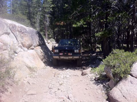 1990 Ford Bronco - 1990 Ford Bronco navigates a challenging off-road path surrounded by trees and large rocks.
