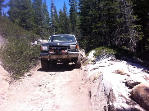 1990 Ford Bronco - A dark blue Ford Bronco navigates a challenging rocky trail surrounded by trees.