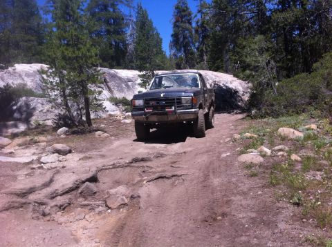 1990 Ford Bronco - 1990 Ford Bronco navigating a challenging off-road trail in a wooded, rocky area.