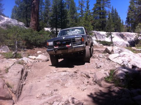 1990 Ford Bronco - A dark blue Ford Bronco navigates a challenging rocky trail under a clear blue sky.