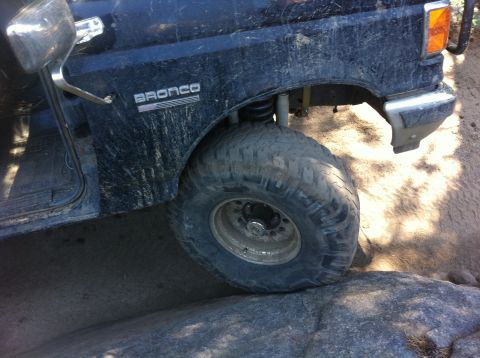 1990 Ford Bronco - The front fender and tire of a dark blue Ford Bronco are shown navigating rough, rocky ground.