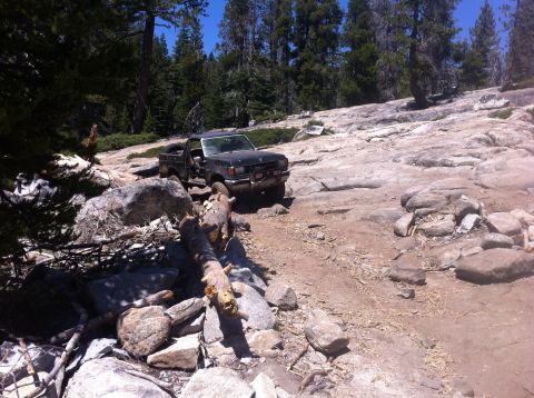 1990 Ford Bronco - A black pickup truck drives over a rocky terrain with trees in the background.