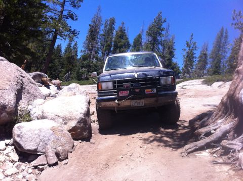 1990 Ford Bronco - A dark blue Ford Bronco drives through a rocky, wooded area on a sunny day.